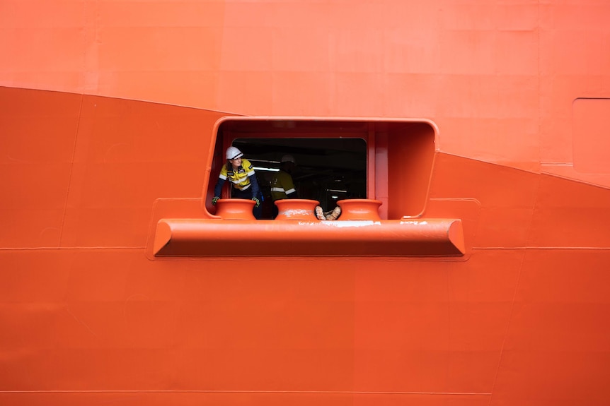 A person wearing a hardhat peers out of a porthole in a large orange ship.