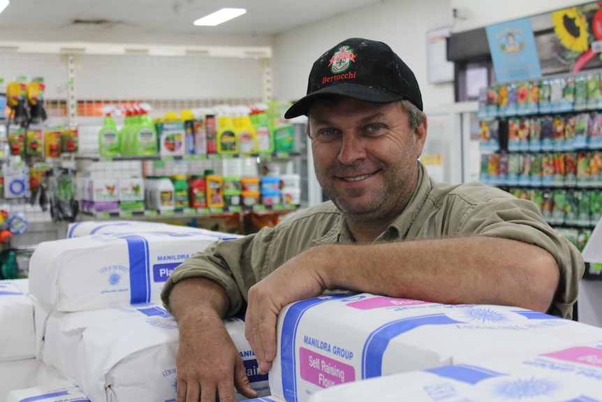 A man with stubble wearing a cap and country-style shirt leans on large packets of flour in a supermarket.