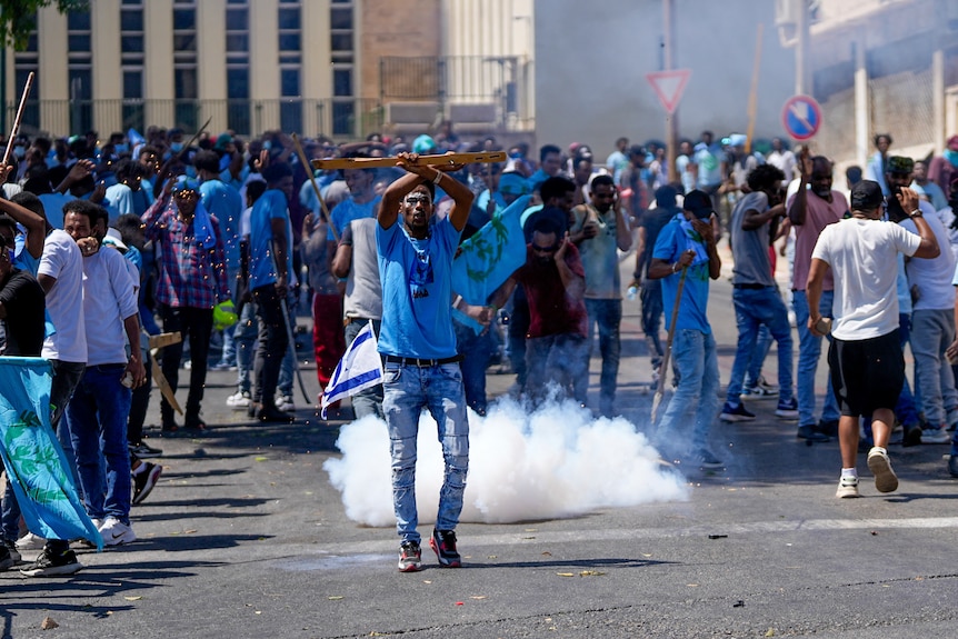A group of protesters in blue shirts stand on a street, in the centre a man with an Israeli flag on his hip, smoke behind him