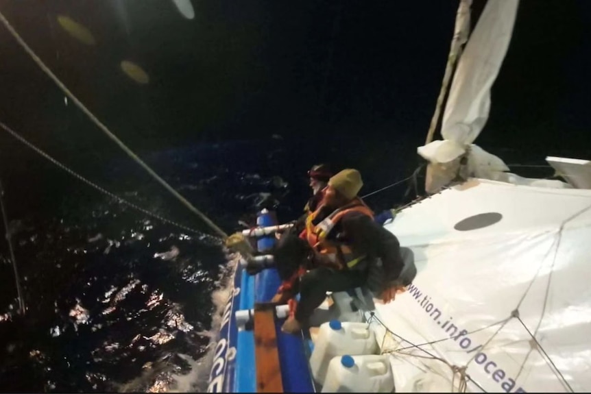Men clinging to a catamaran at night while a rescue operation takes place.