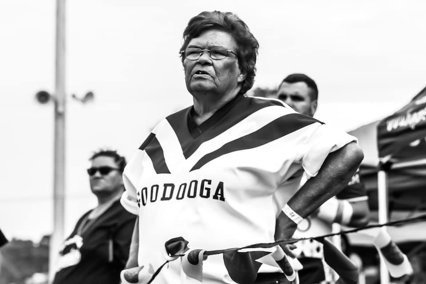 A black and white image shows a woman in a 'Goodooga' NRL jersey with her hands on her hips