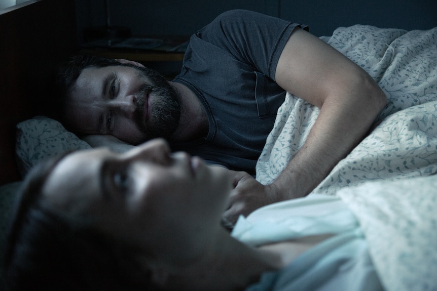Man and woman lying in a dark, cooly lit room in bed. Man looks intensely at woman in foreground and pulls sheets up around him.
