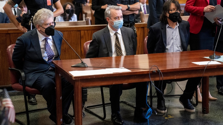 Three men wearing face masks sit at the front of a courtroom.