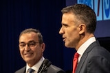 Two men stand in front of microphones, one is facing the camera and one is side on