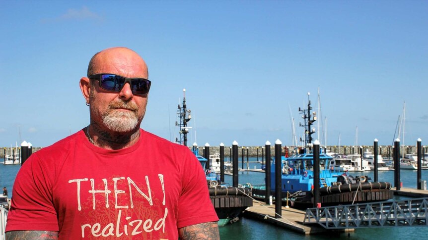 Scott Porter, heavily tattooed, stands by a marina. He is wearing a red shirt and large sunglasses.