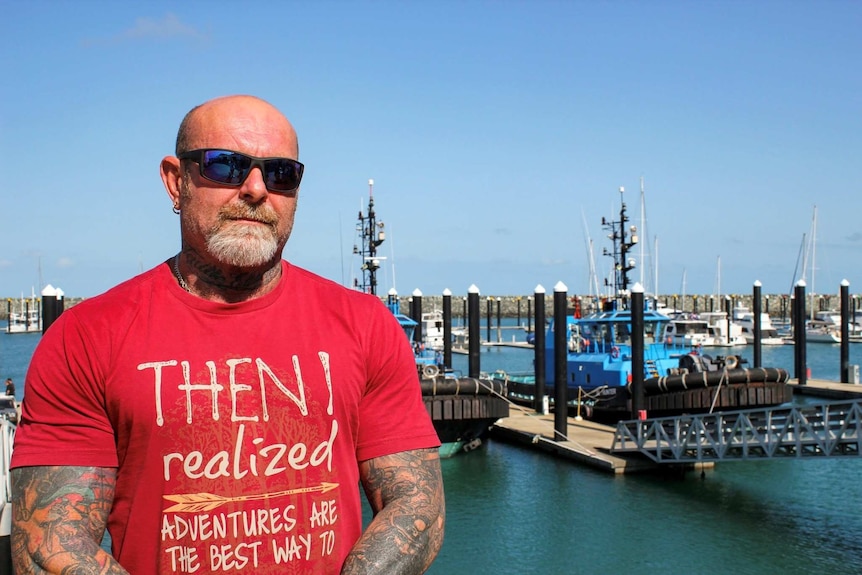 Scott Porter, heavily tattooed, stands by a marina. He is wearing a red shirt and large sunglasses.