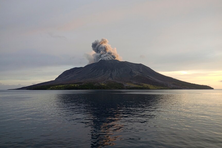 A grey cloud rises from a volcanic islands sitting in the middle of a dark sea.