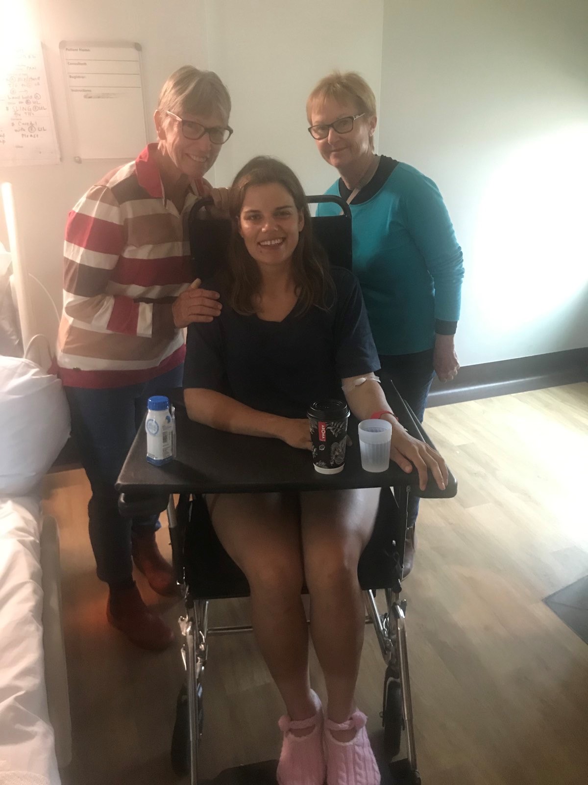 Young woman in wheelchair with two older women standing either side in hospital room