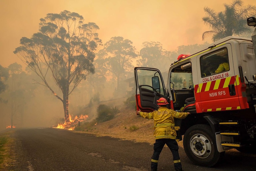 A fire truck and a firefighter see a blazing bushfire burning near a home