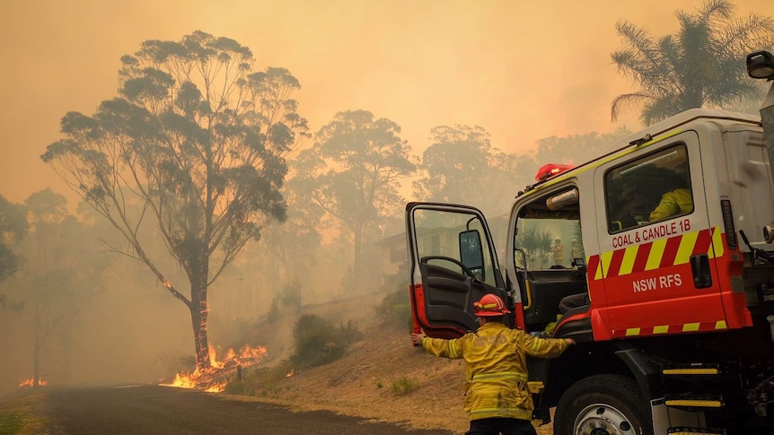 A fire truck and a firefighter see a blazing bushfire burning near a home
