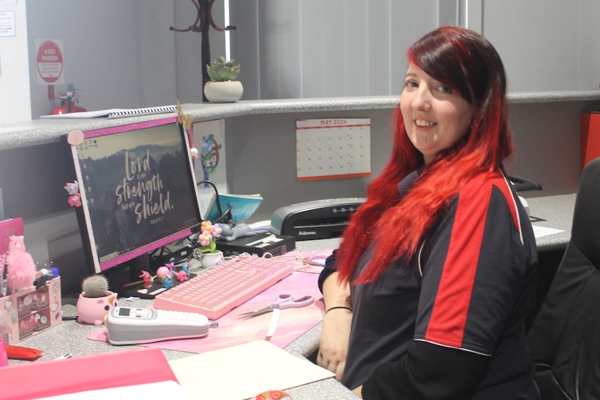 A woman with black and red hair sits at an office desk with a pink keyboard.