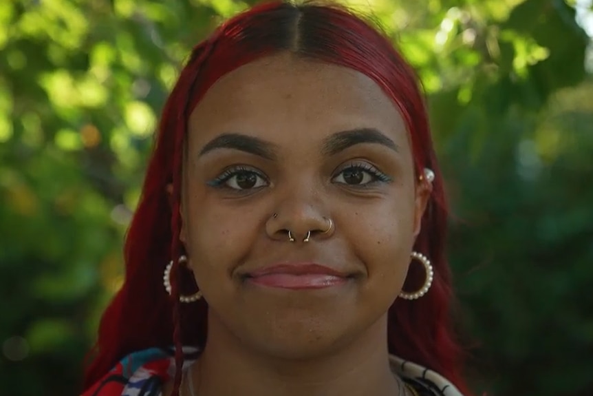 A young woman with brown skin and red hair smiles at the camera. She has gold earrings with pearls and a gold nose ring.