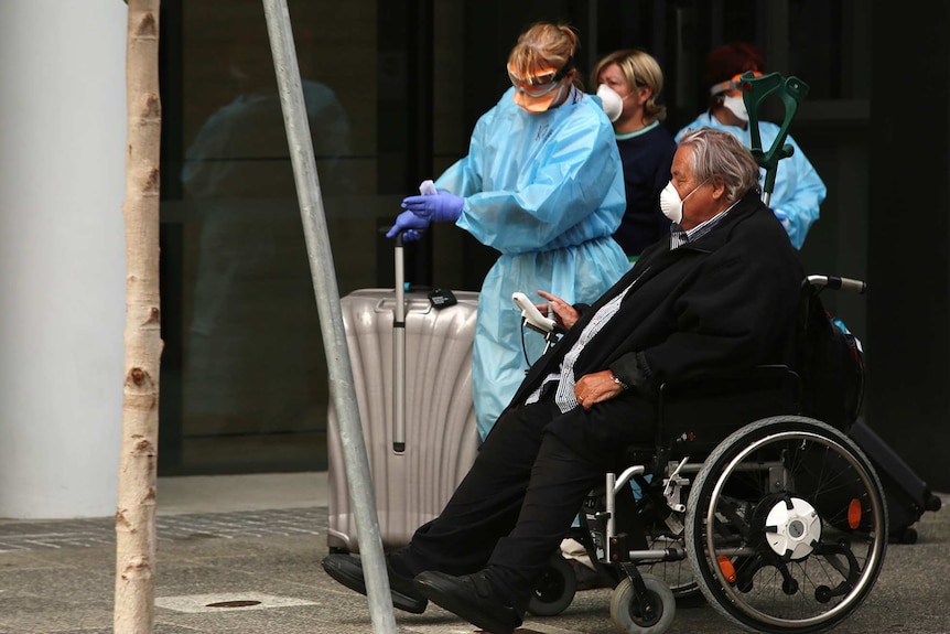 A man in a wheelchair wearing a facemask is escorted along a footpath by a woman wearing full-body protective clothing.