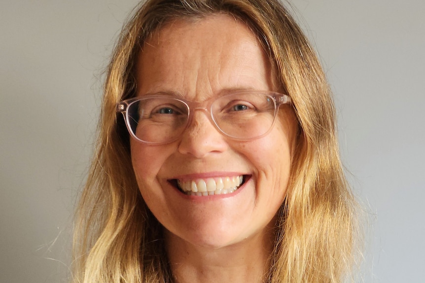 Portrait of Rachel Schofield with long blonde hair and a wide smile, wearing a royal blue shirt and reading glasses. 