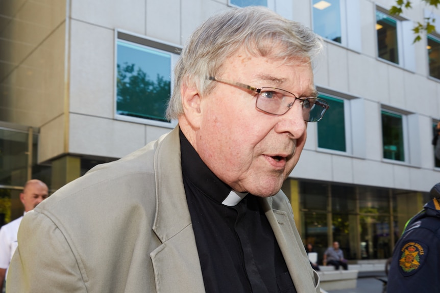 An older man wears a grey suit jacket and black priest's shirt. 