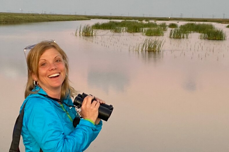 A lady holding binoculars, wearing a blue jumper, in front of a pink-toned lagoon. 