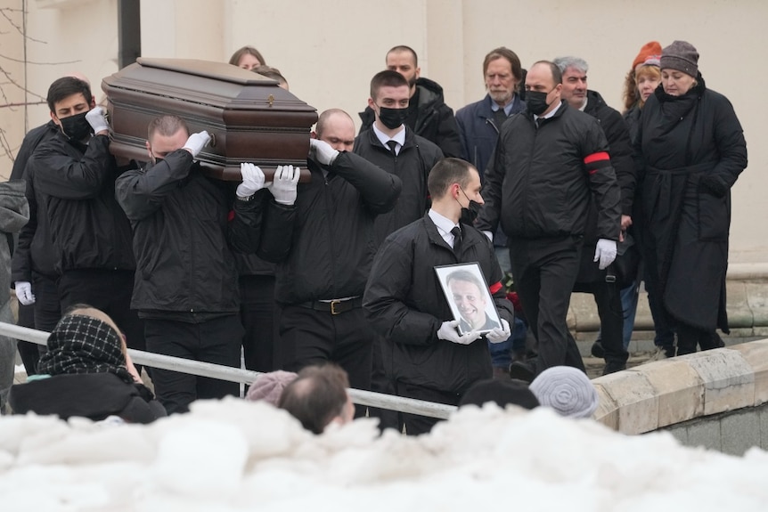 A large group of people walking, some carrying a coffin, some walking behind it, and one in front of it carrying a framed photo