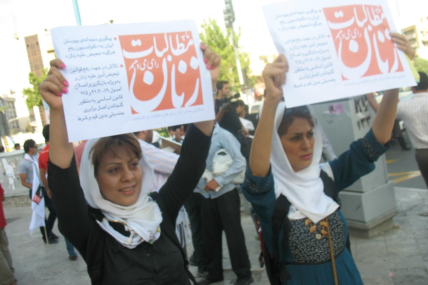 Two women wearing white scarves over their hair hold placards with red graphic and Iranian script