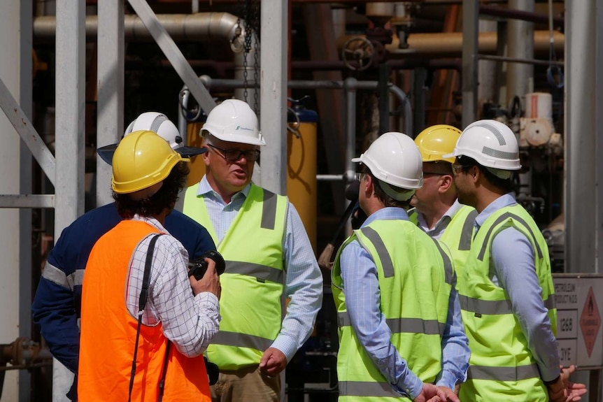 A group in high vis vests and hard hats stand at an oil refinery in a group talking.