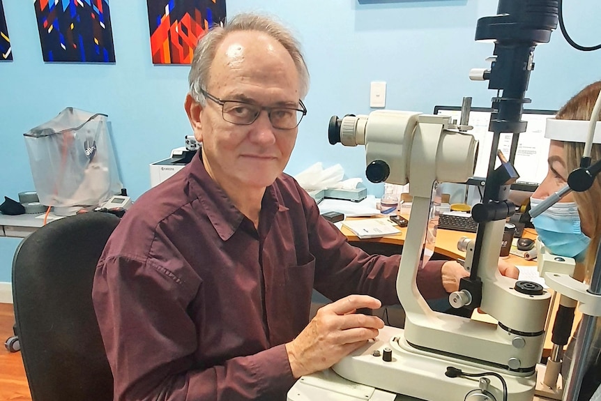 A man in a button up shirt smiles at the camera with eye testing machines in front of him