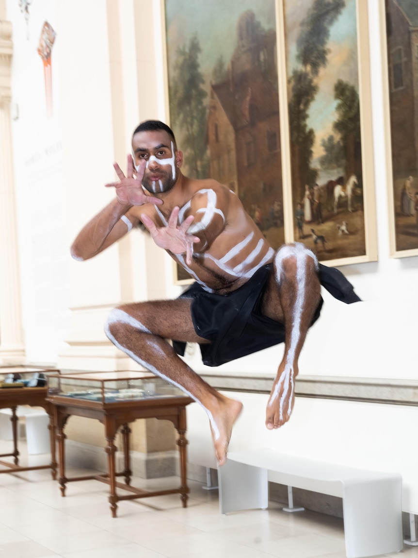 Jack Collard leaps in the air in front of oil paintings in Brussels.  He is wearing traditional clothing and body paint.
