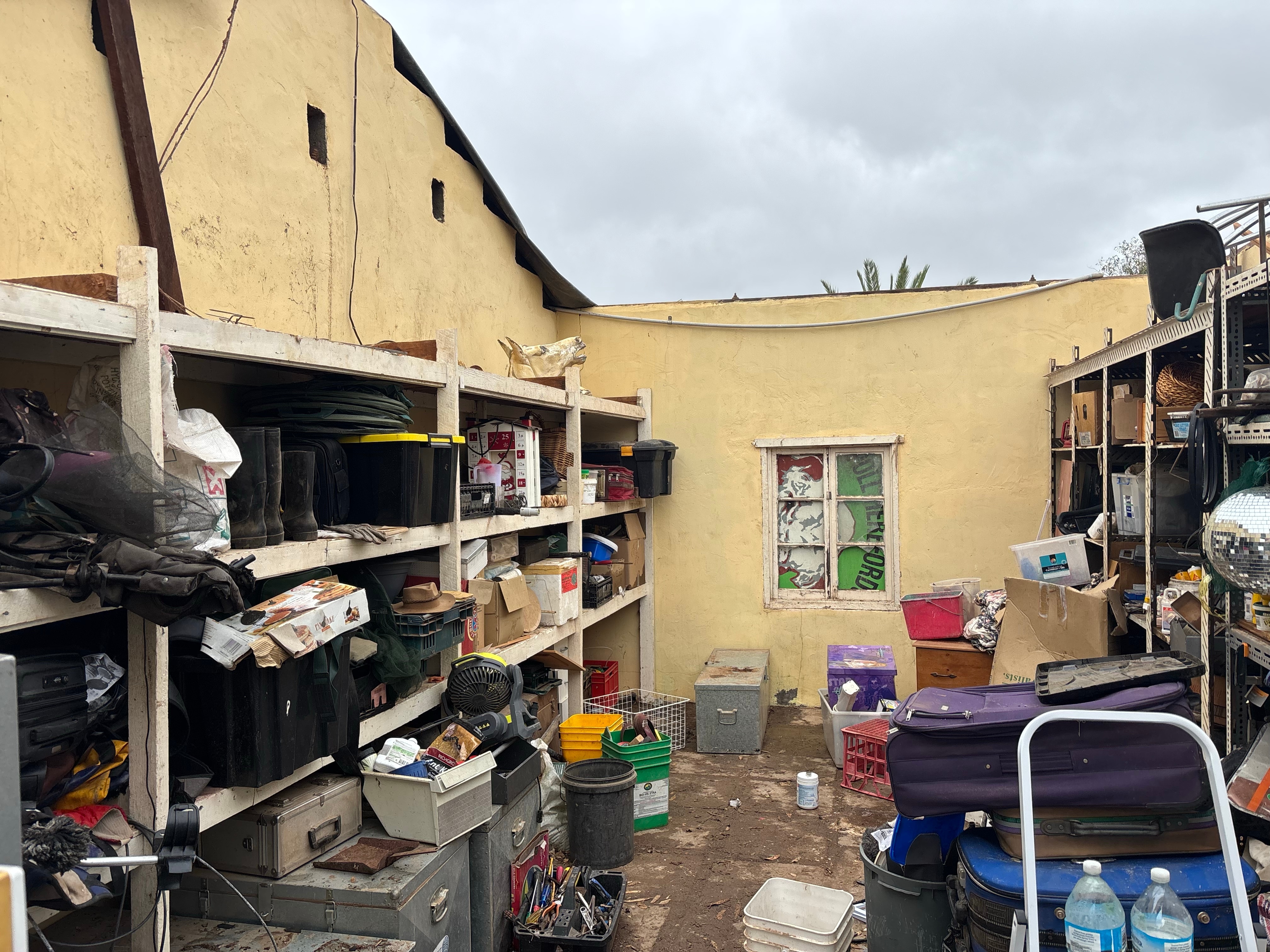 A view inside a yellow walled store room with shelves full of stuff, but no roof.
