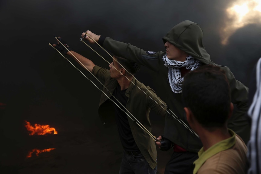 Palestinian protesters draw back slingshots to hurl stones at Israeli troops in Gaza with smoke in the background.