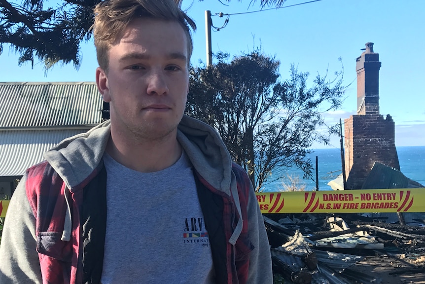 A young man in a grey shirt and hoodie standing in front of a house destroyed by fire.