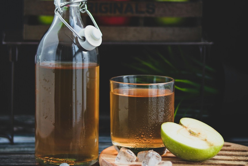 A glass of apple cider vinegar next to a glass bottle of the same liquid and half an apple.