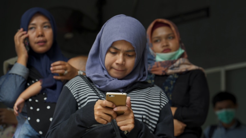 A woman in a headscarf stands with two other women, as she looks down in concentration at her phone