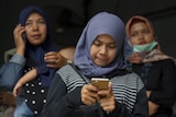 A woman in a headscarf stands with two other women, as she looks down in concentration at her phone