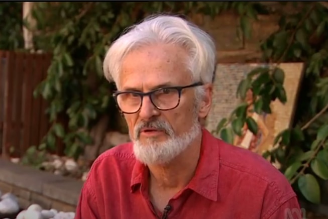 A grey-haired man  in a red shirt with grey hair sitting on a chair.