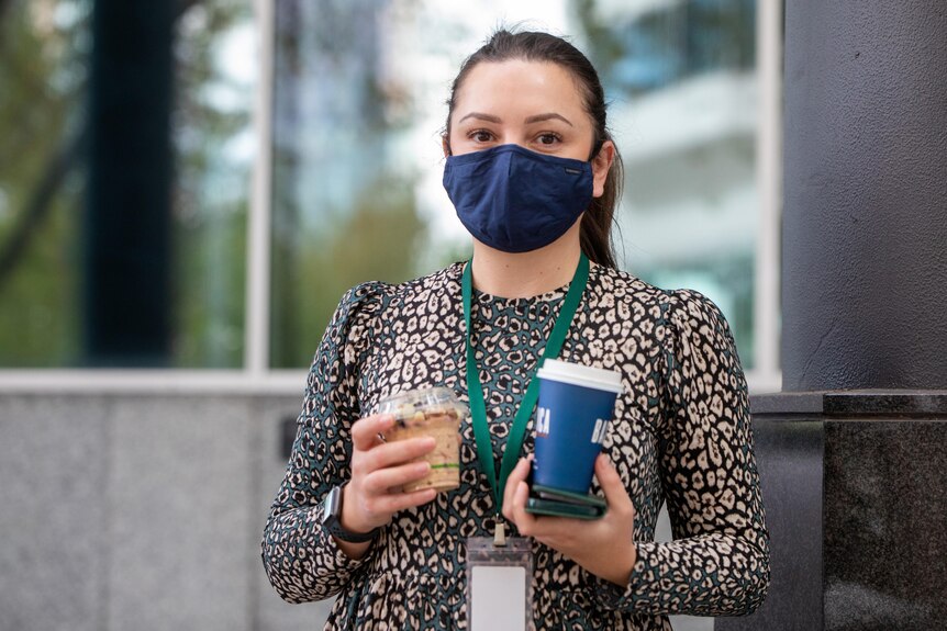 A mid-shot of a woman wearing a face mask carrying two takeaway coffees.