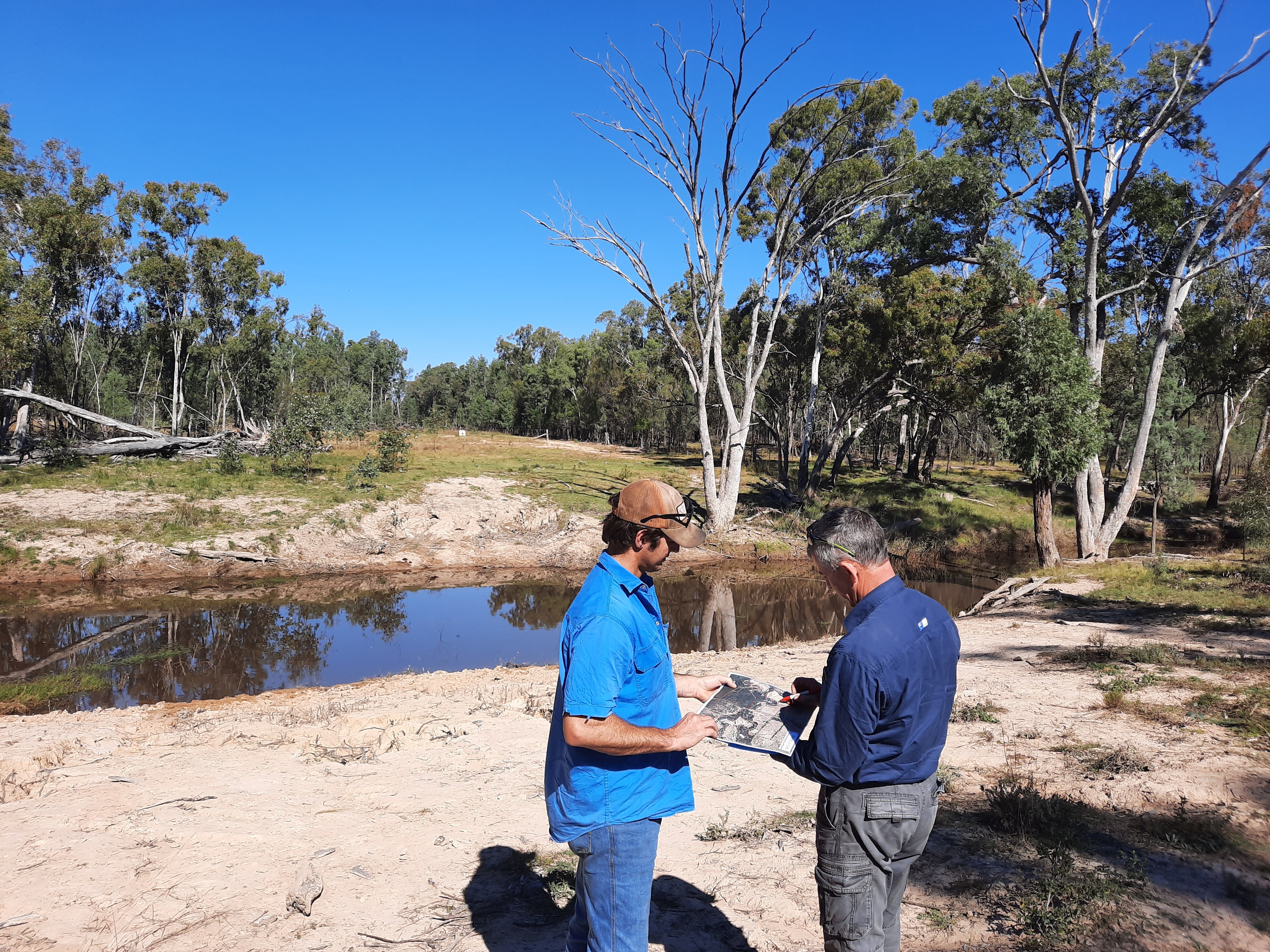 Two men standing on a river bank looking at a map