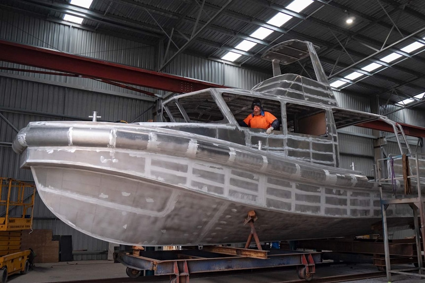 A stainless steel boat being built in a large shed with a worker in the cabin