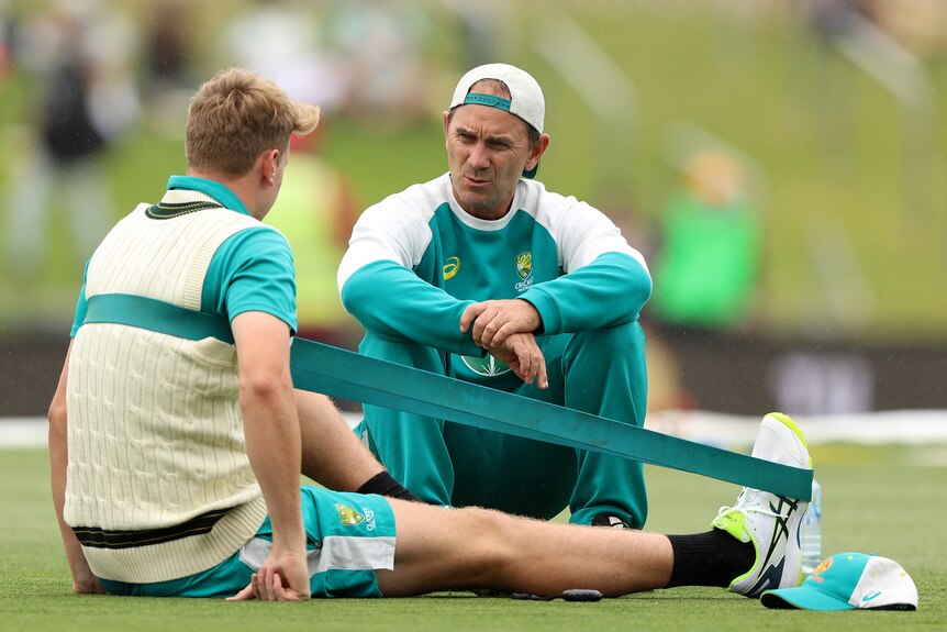 Justin Langer, with cap on backwards, sits on the grass and chats with Cameron Green