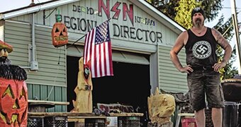 A man wearing singlet with swastika stands in front of warehouse with American flag hanging from it.