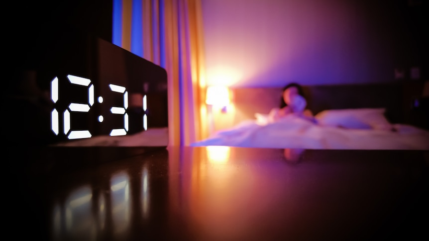 A clock reading 12:31 in the foreground with a woman in bed in the background