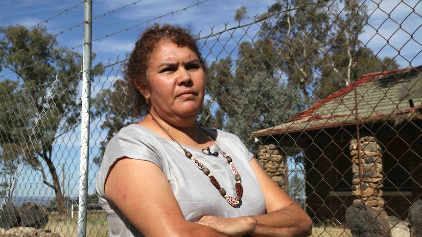 A woman stands in front of an old stone cottage and a barbed wire fence.