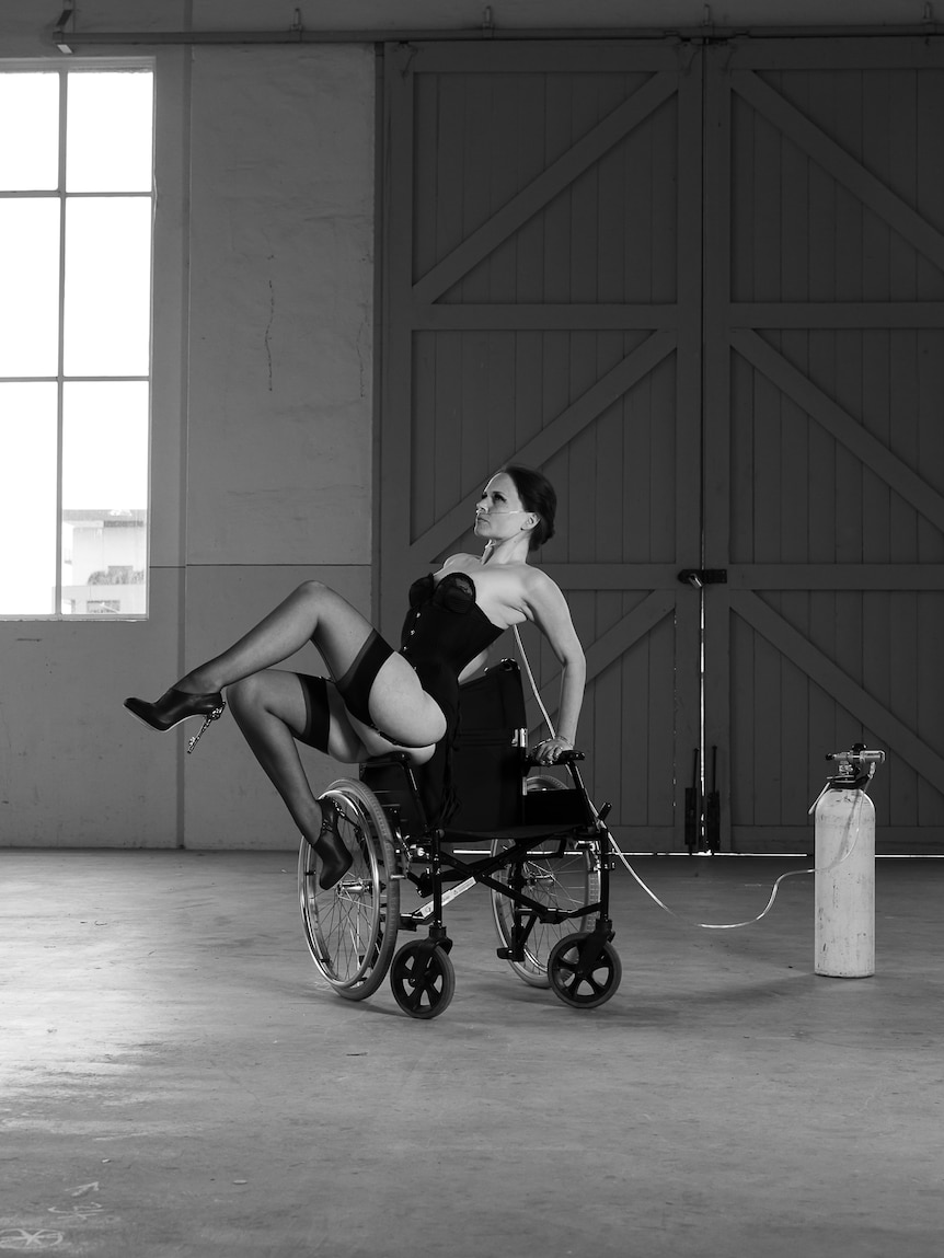 A woman in her mid 30s in lingerie holds her body up from a wheelchair, hooked up to oxygen