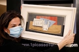 A woman wearing a face mask holds up a framed painting with red, brown and white colours.