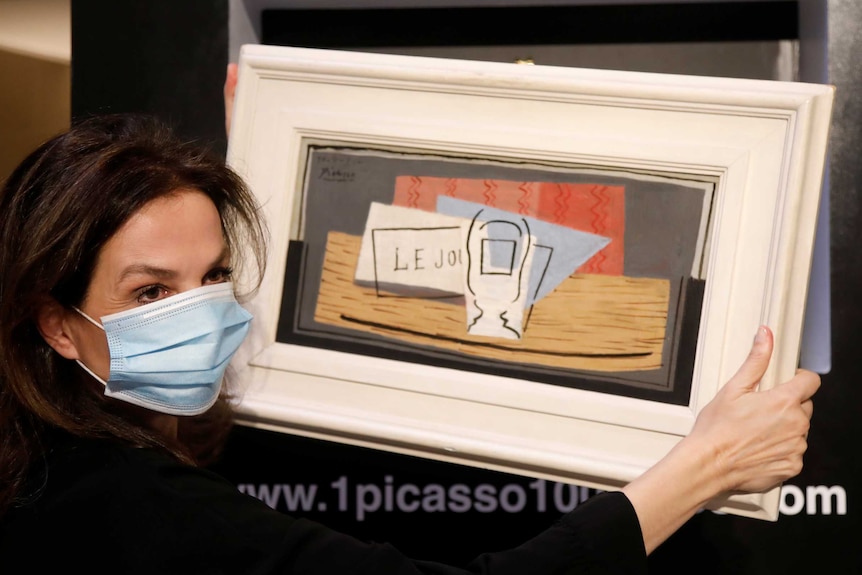 A woman wearing a face mask holds up a framed painting with red, brown and white colours.