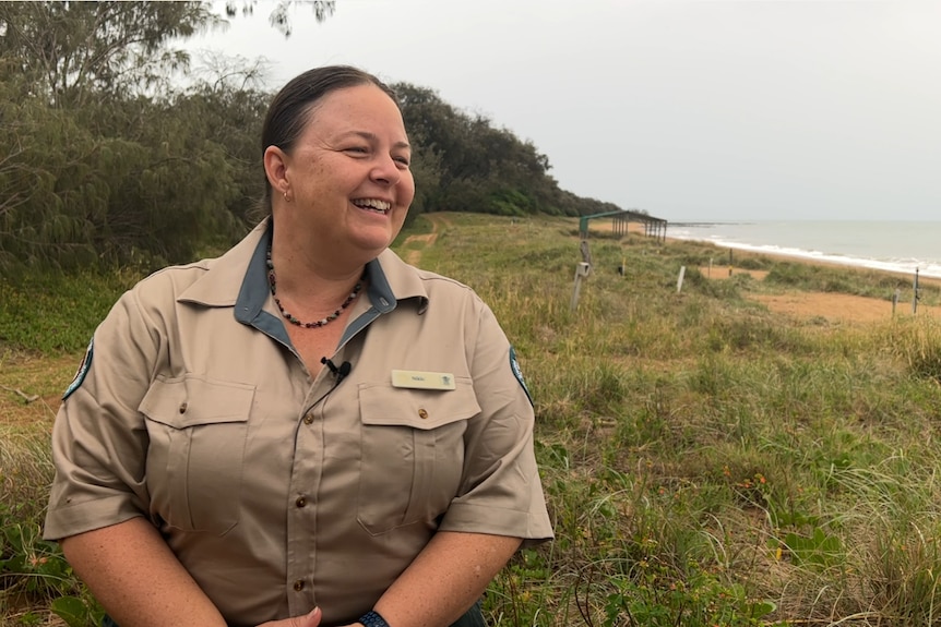 A woman wearing khaki sits on a stump on a sand dune with the ocean and beach in the background. 