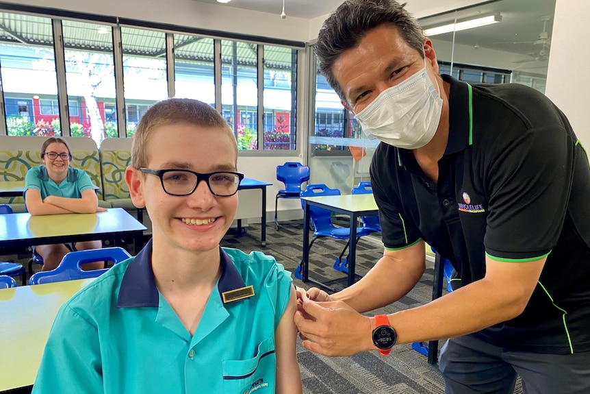 A boy smiles at the camera while a man injects him