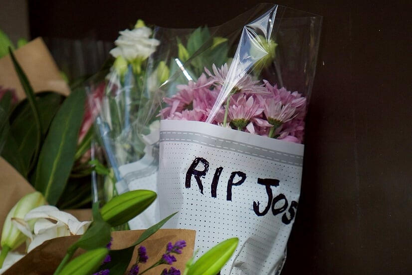 A bouquet of pink flowers wrapped in paper reading 'RIP Josh' is laid amongst a row of tributes.