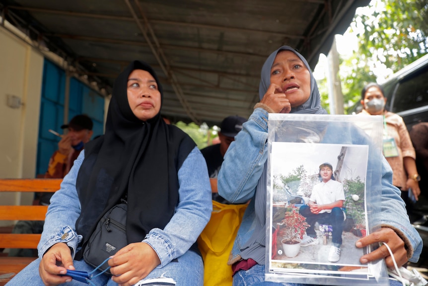 Two women sit on a bench, with one holding a laminated photo of an adult son.
