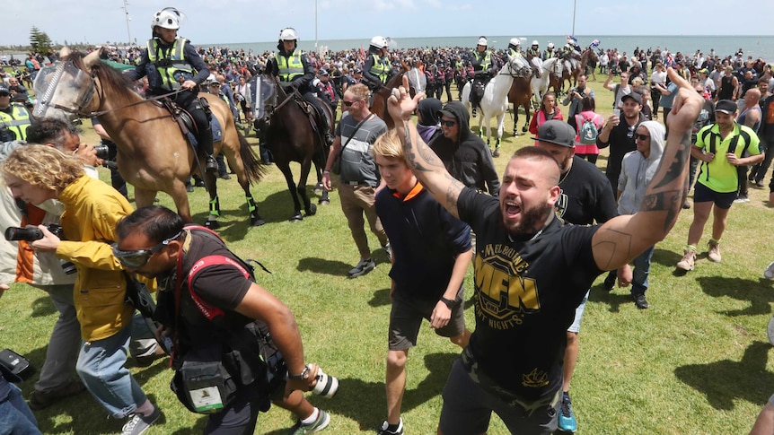 A tattooed man raises both arms in the air, pointing to the sky, while walking through a crowd. Police on horseback watch.