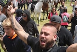 A tattooed man raises both arms in the air, pointing to the sky, while walking through a crowd. Police on horseback watch.