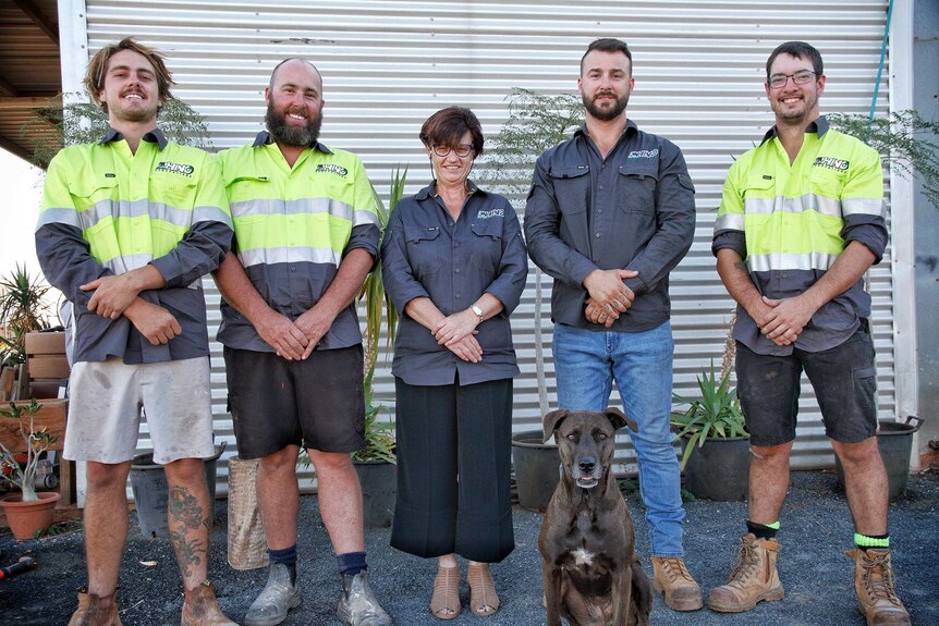 Five people stand outdoors wearing work clothes and hi-vis shirts posing for a photo with a dog.