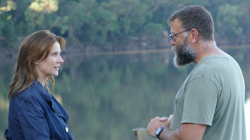 Rachel Griffiths is pictured side-on, speaking with a man who has his hands clasped. There is a river behind them. 